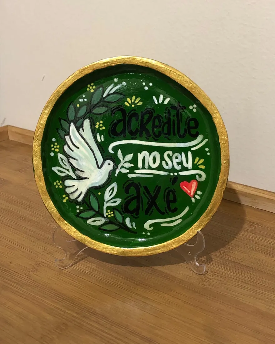 Prato decorativo pintado à mão num suporte, verde profundo com borda dourada, mostrando uma pomba branca com ramos de oliveira e a frase "Acredite no seu axé" em letras feitas à mão.