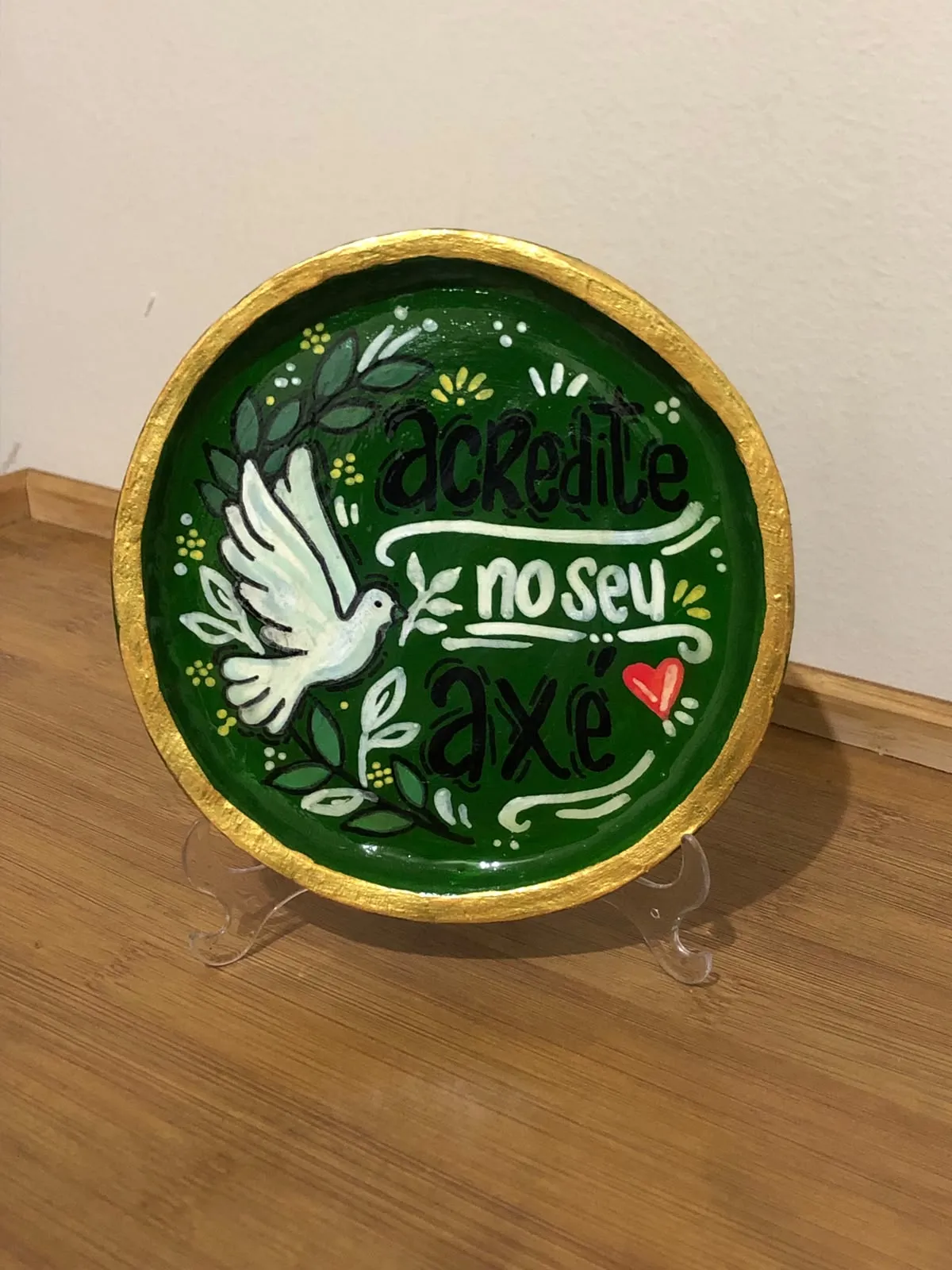 Prato decorativo pintado à mão num suporte, verde profundo com borda dourada, mostrando uma pomba branca com ramos de oliveira e a frase "Acredite no seu axé" em letras feitas à mão.