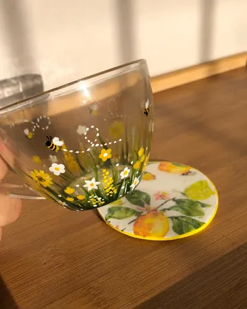 Clear glass mug hand-painted with daisies, yellow wildflowers, grass and small bees, held up in sunlight next to a lemon-print coaster.