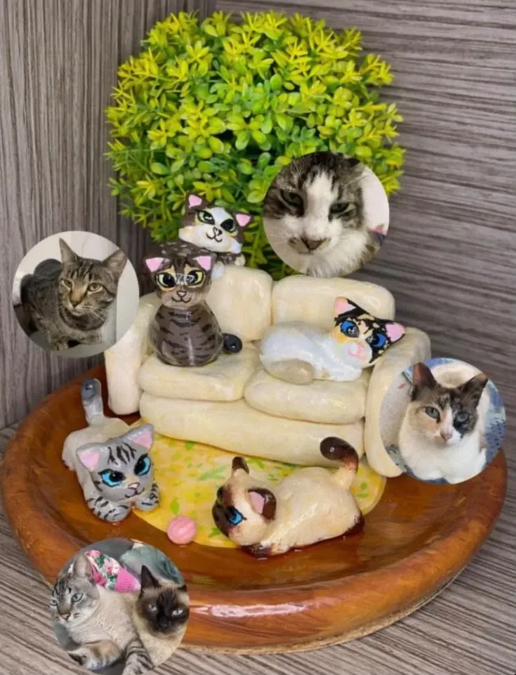 Miniatura em argila de cinco gatos sobre e em volta de um sofá creme numa base de madeira, ao lado das fotos dos gatos reais que serviram de modelo.