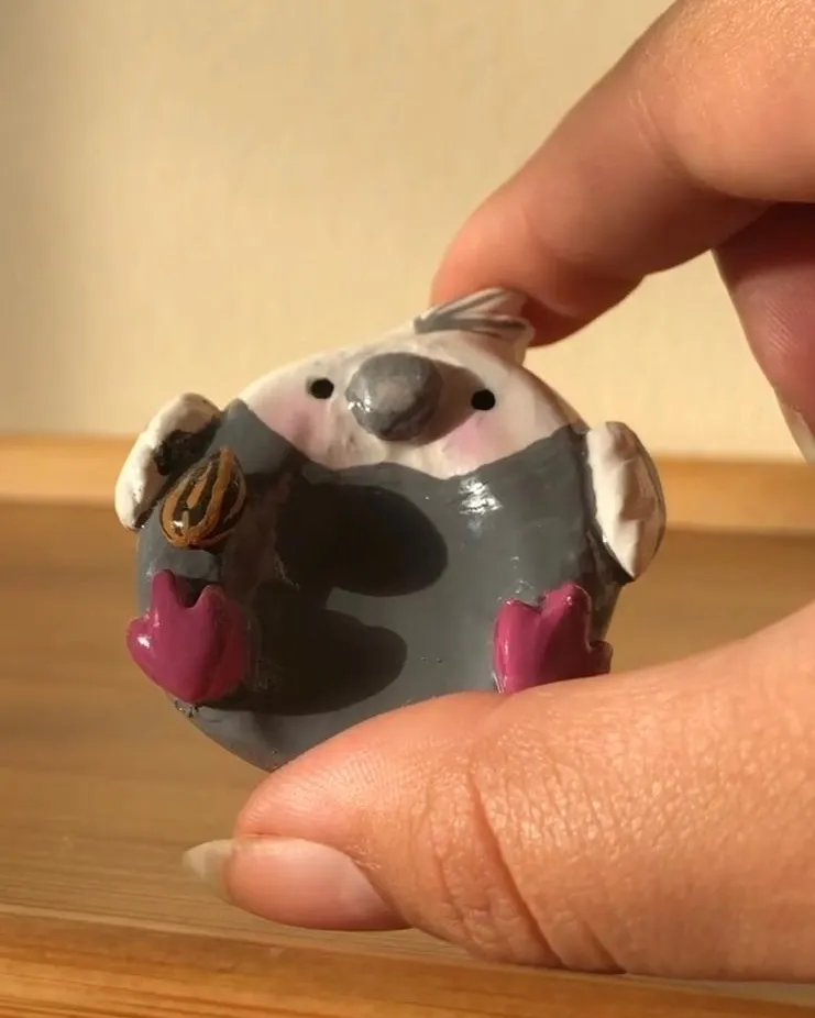 Miniatura de calopsita cinza esculpida à mão, com bochechas rosadas, segurando uma semente de girassol entre os dedos de uma mão.