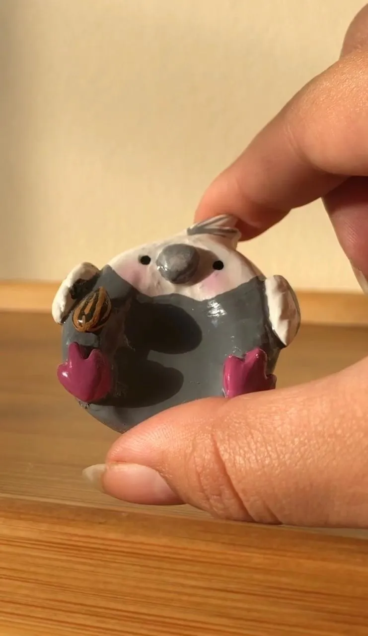 Miniatura de calopsita cinza esculpida à mão, com bochechas rosadas, segurando uma semente de girassol entre os dedos de uma mão.
