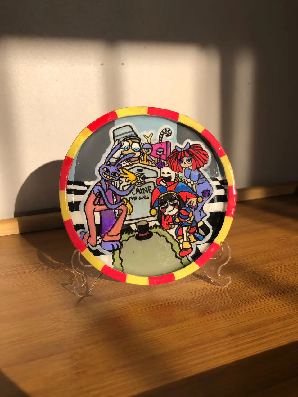 Prato decorativo pintado à mão num suporte, com borda vermelha e amarela, mostrando os personagens de The Amazing Digital Circus reunidos em volta de uma lápide.