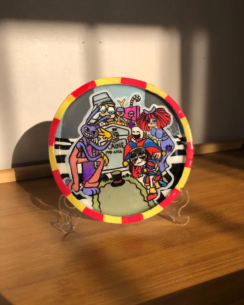 Prato decorativo pintado à mão num suporte, com borda vermelha e amarela, mostrando os personagens de The Amazing Digital Circus reunidos em volta de uma lápide.