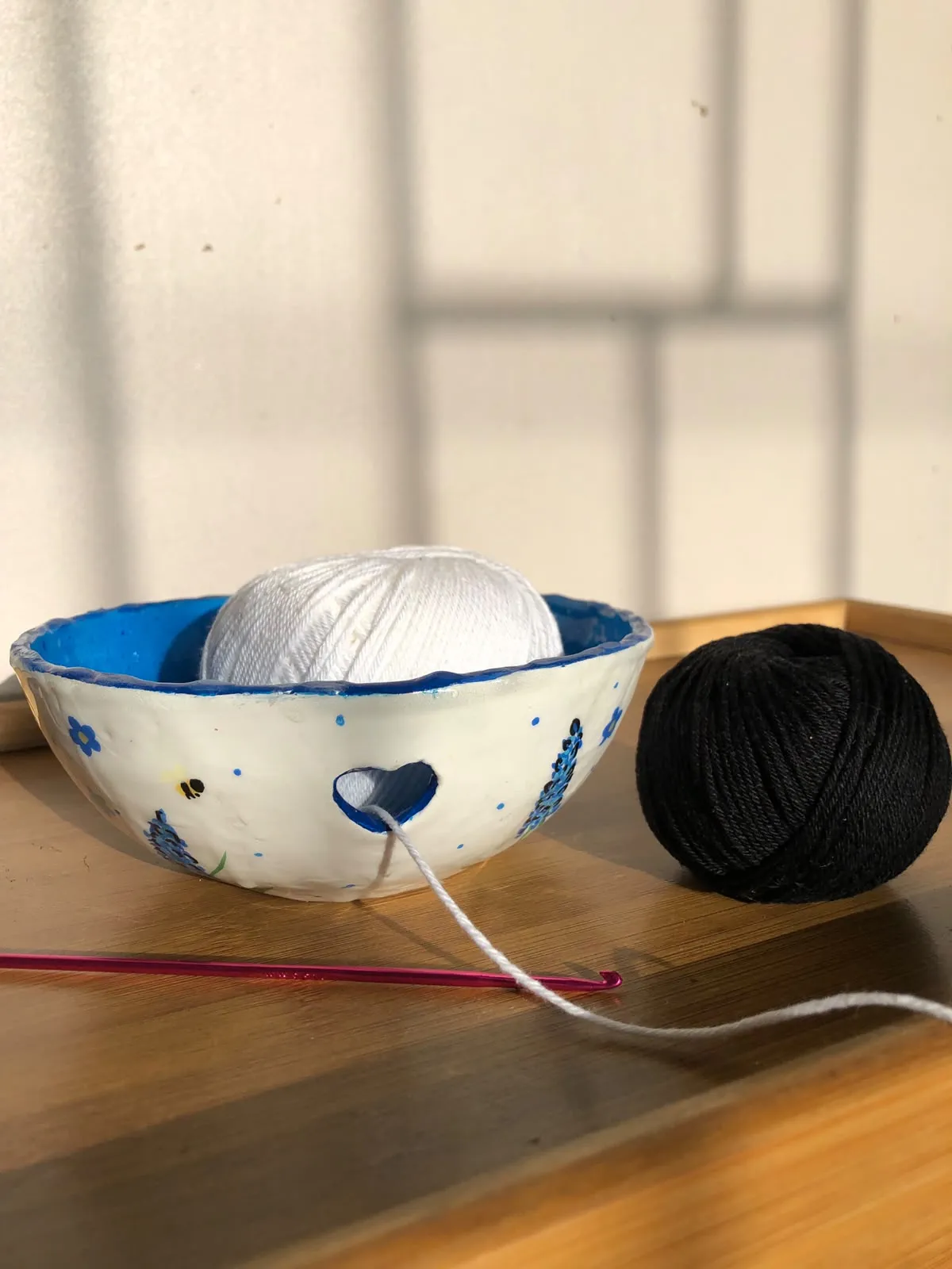 Tigela de lã branca esculpida à mão com um recorte em formato de coração, pintada com lavandas e abelhas, com um novelo branco dentro, ao lado de um novelo preto e uma agulha de crochê rosa.