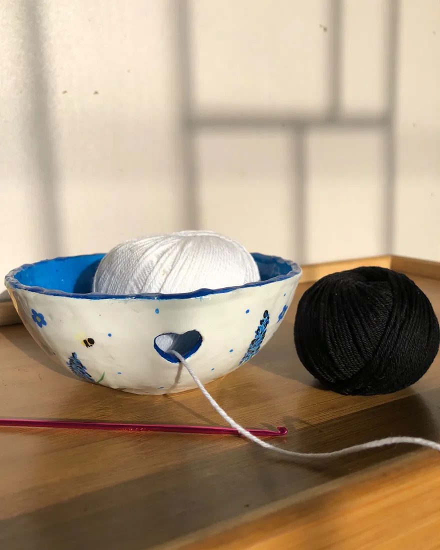 Tigela de lã branca esculpida à mão com um recorte em formato de coração, pintada com lavandas e abelhas, com um novelo branco dentro, ao lado de um novelo preto e uma agulha de crochê rosa.