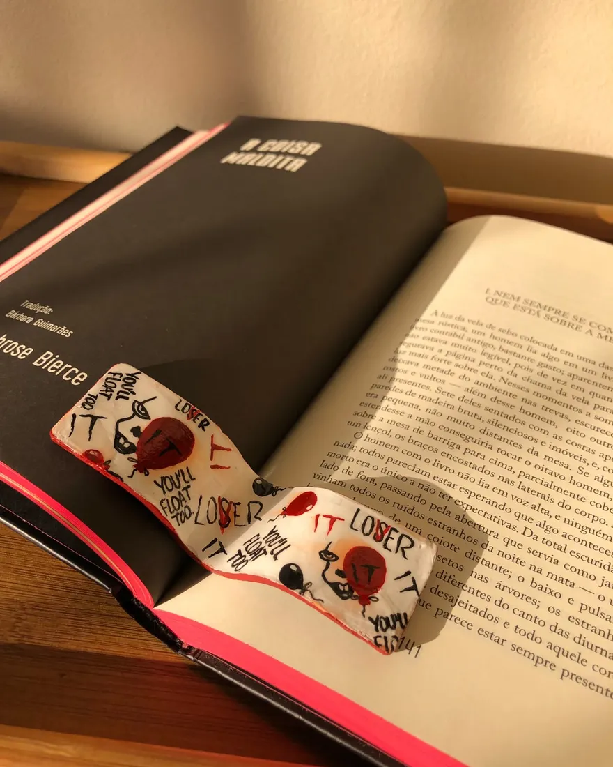 Marcador de páginas branco pintado à mão com balões vermelhos e as palavras "You'll float too" e "LOSER", sobre as páginas de um livro aberto.