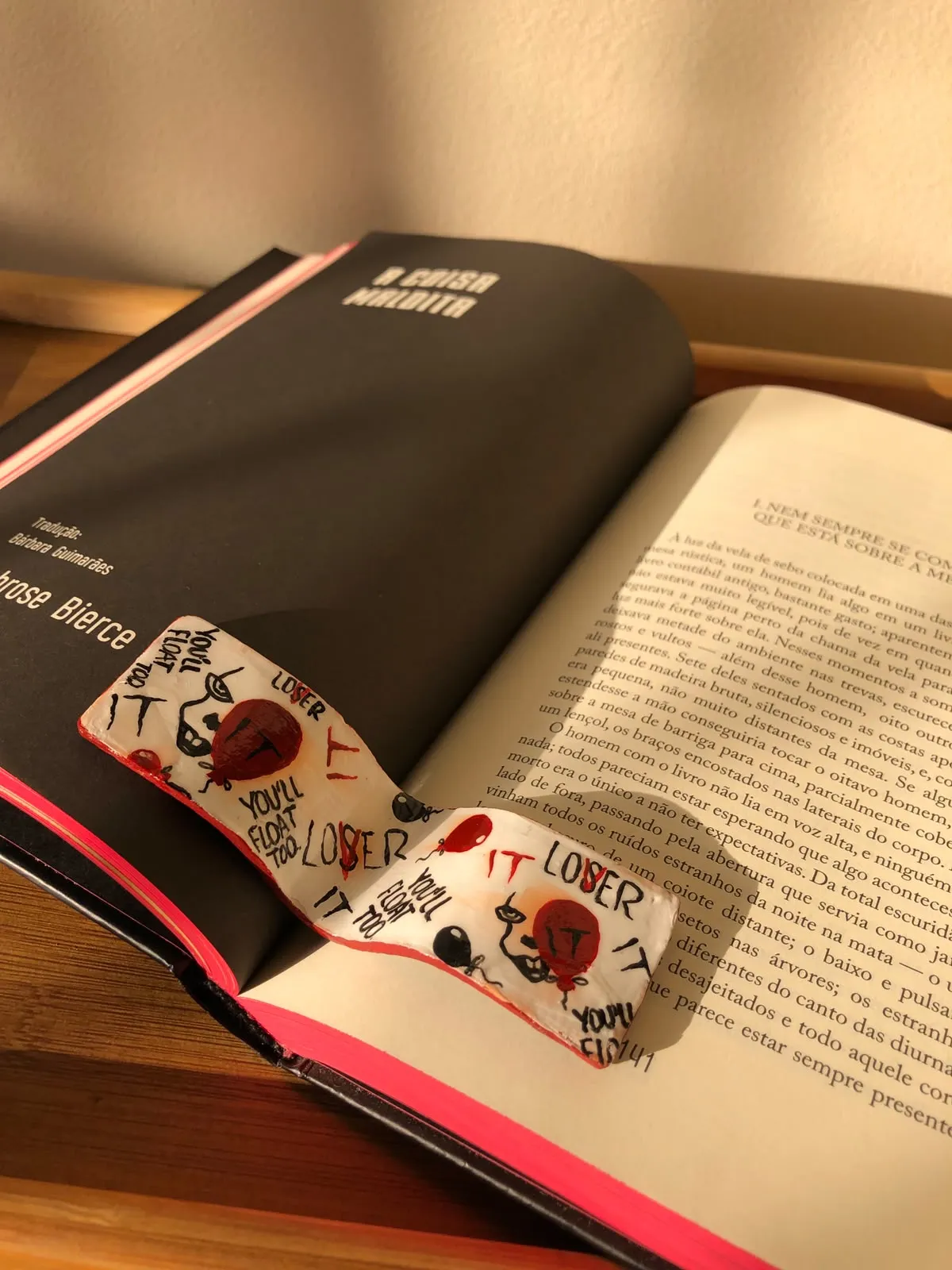 Marcador de páginas branco pintado à mão com balões vermelhos e as palavras "You'll float too" e "LOSER", sobre as páginas de um livro aberto.