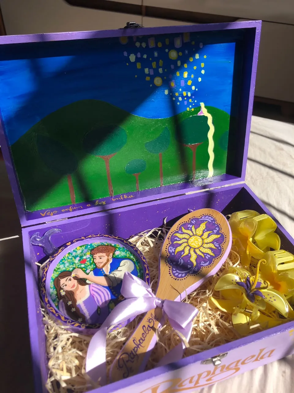 Caixa de madeira roxa aberta, pintada com a cena das lanternas de Enrolados na tampa, contendo uma escova pintada, um espelho de mão com Rapunzel e Flynn e presilhas amarelas.