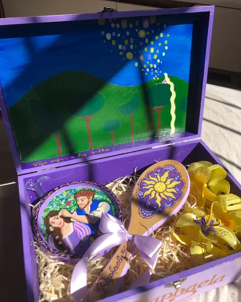 Caixa de madeira roxa aberta, pintada com a cena das lanternas de Enrolados na tampa, contendo uma escova pintada, um espelho de mão com Rapunzel e Flynn e presilhas amarelas.