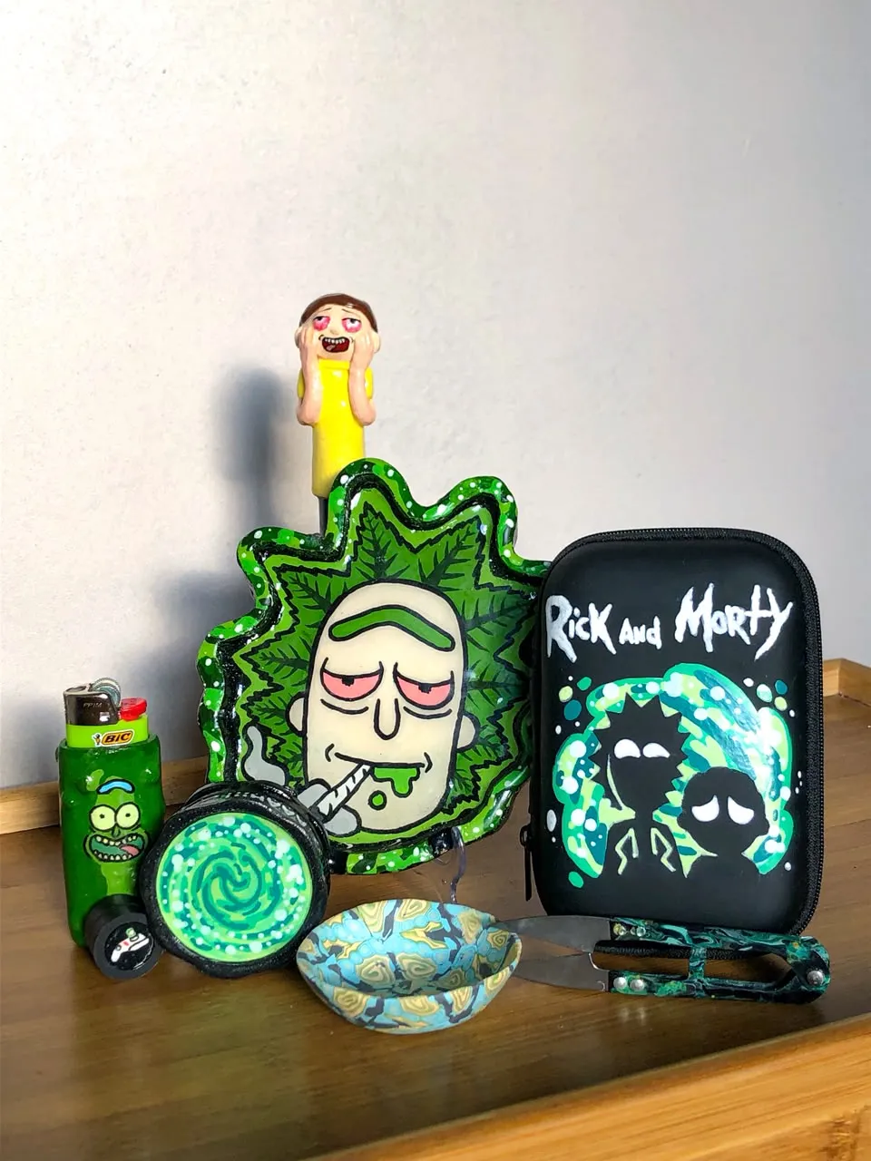 Kit temático de Rick and Morty pintado à mão sobre uma mesa de madeira, bandeja verde do Rick, dichavador com portal, isqueiro do Pickle Rick, estojo preto pintado e um pequeno boneco do Morty.