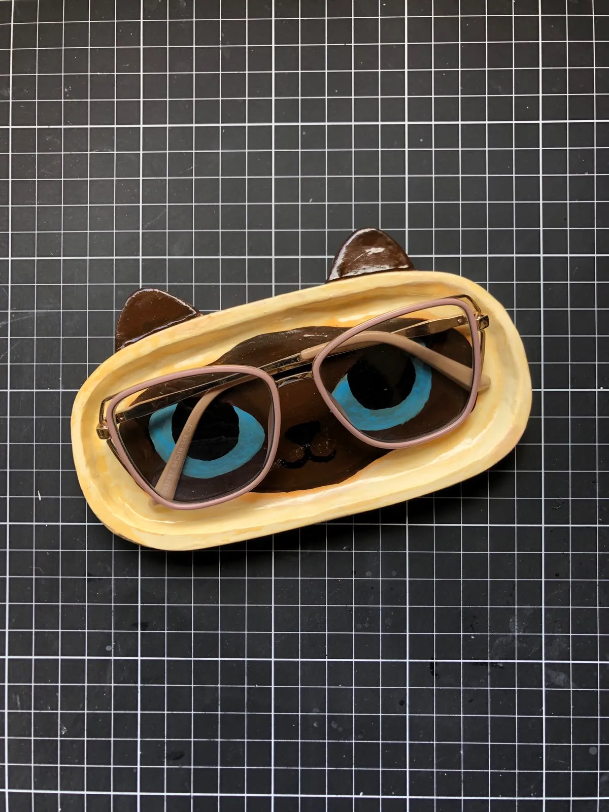 Oval hand-sculpted trinket tray painted as a Siamese cat face with blue eyes and brown ears, holding a pair of glasses, on a black cutting mat.