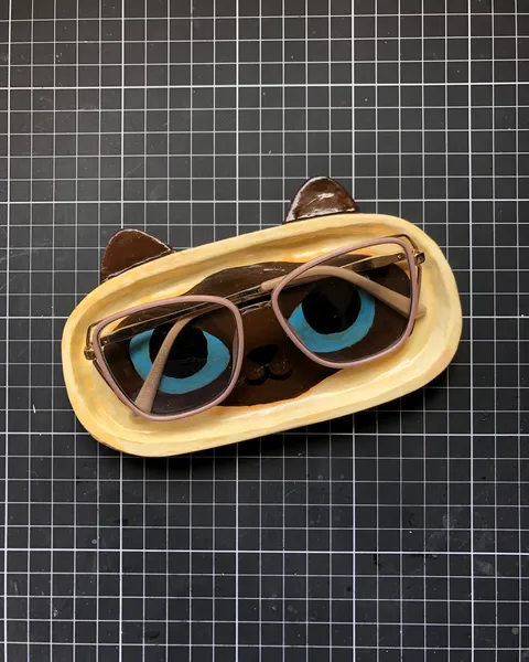 Bandejinha oval esculpida à mão e pintada como o rosto de um gato siamês com olhos azuis e orelhas marrons, com um par de óculos apoiado, sobre uma base de corte preta.