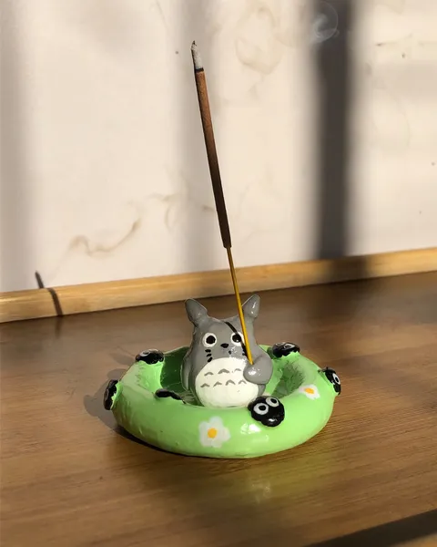 Hand-sculpted incense holder shaped like Totoro sitting in a green pool float with soot sprites, holding a burning incense stick, on a wooden table in sunlight.
