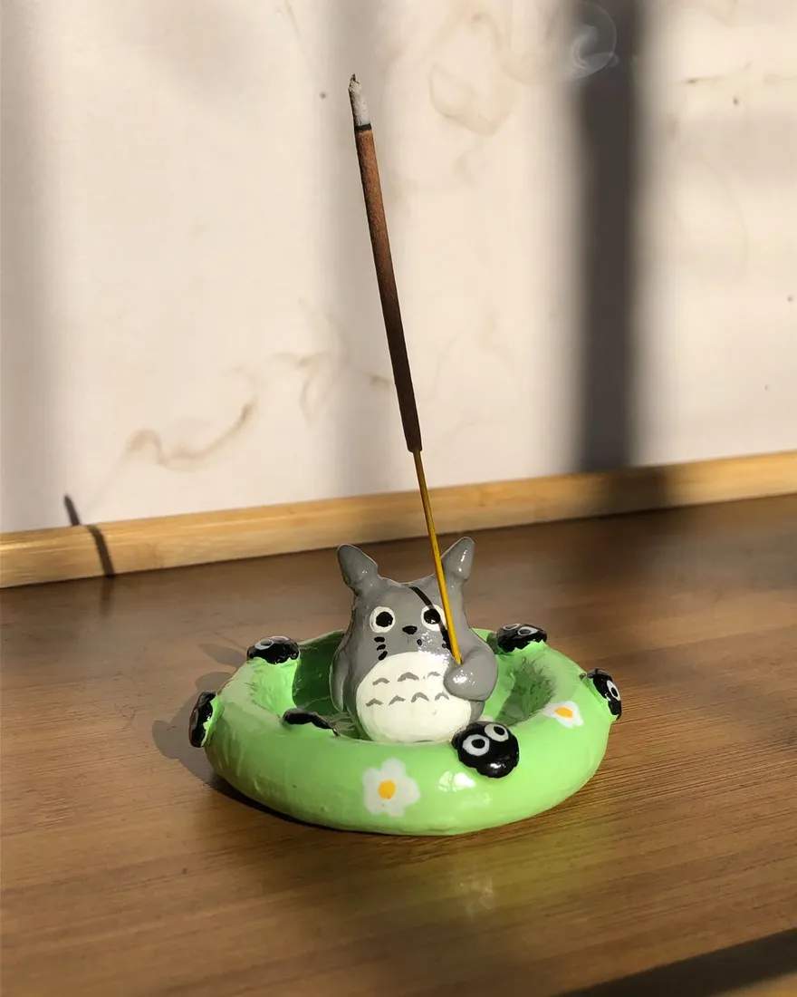Incensário esculpido à mão em formato de Totoro sentado numa boia verde com fuligens, segurando um incenso aceso, sobre uma mesa de madeira ao sol.