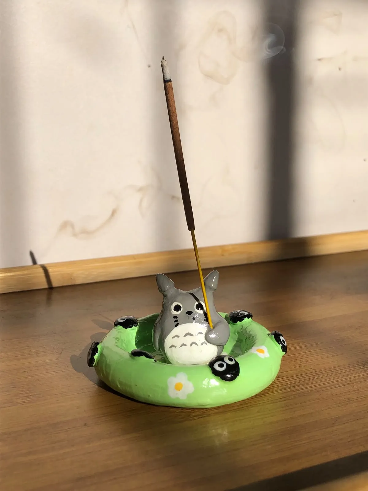 Incensário esculpido à mão em formato de Totoro sentado numa boia verde com fuligens, segurando um incenso aceso, sobre uma mesa de madeira ao sol.
