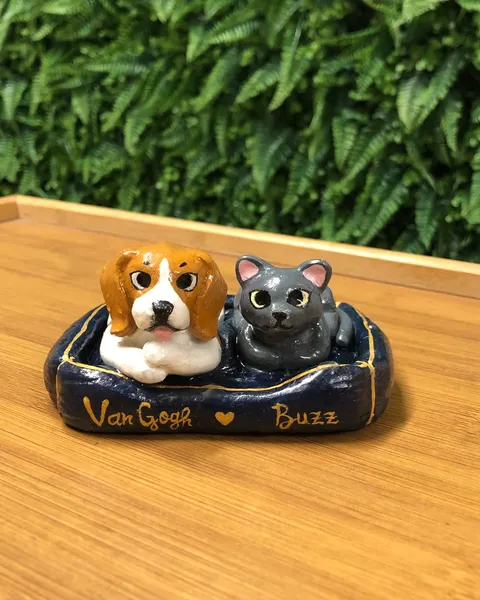 Miniatura em argila de um beagle e um gato cinza deitados juntos numa caminha azul-marinho com os nomes Van Gogh e Buzz pintados em dourado.